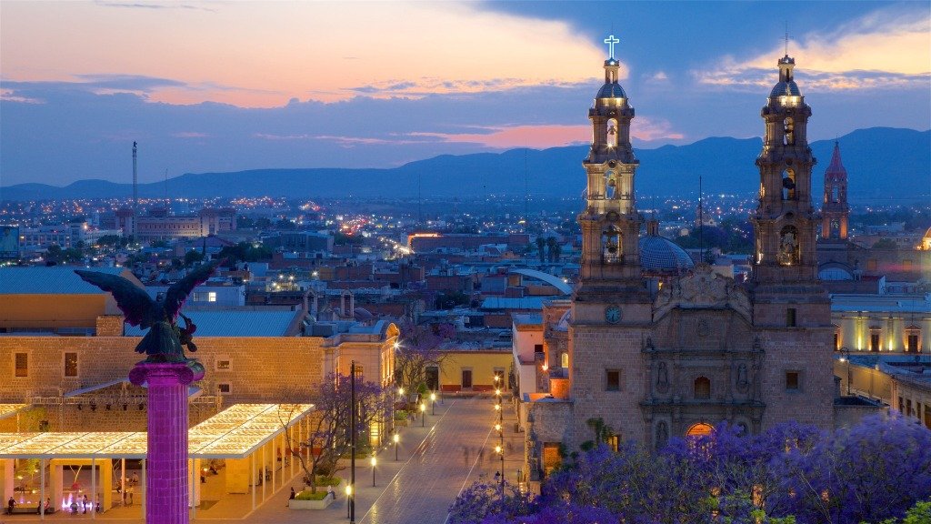 Panorámica crepuscular de Aguascalientes: Plaza de la Patria, Exedra monumental iluminada, Catedral y jacarandas en flor